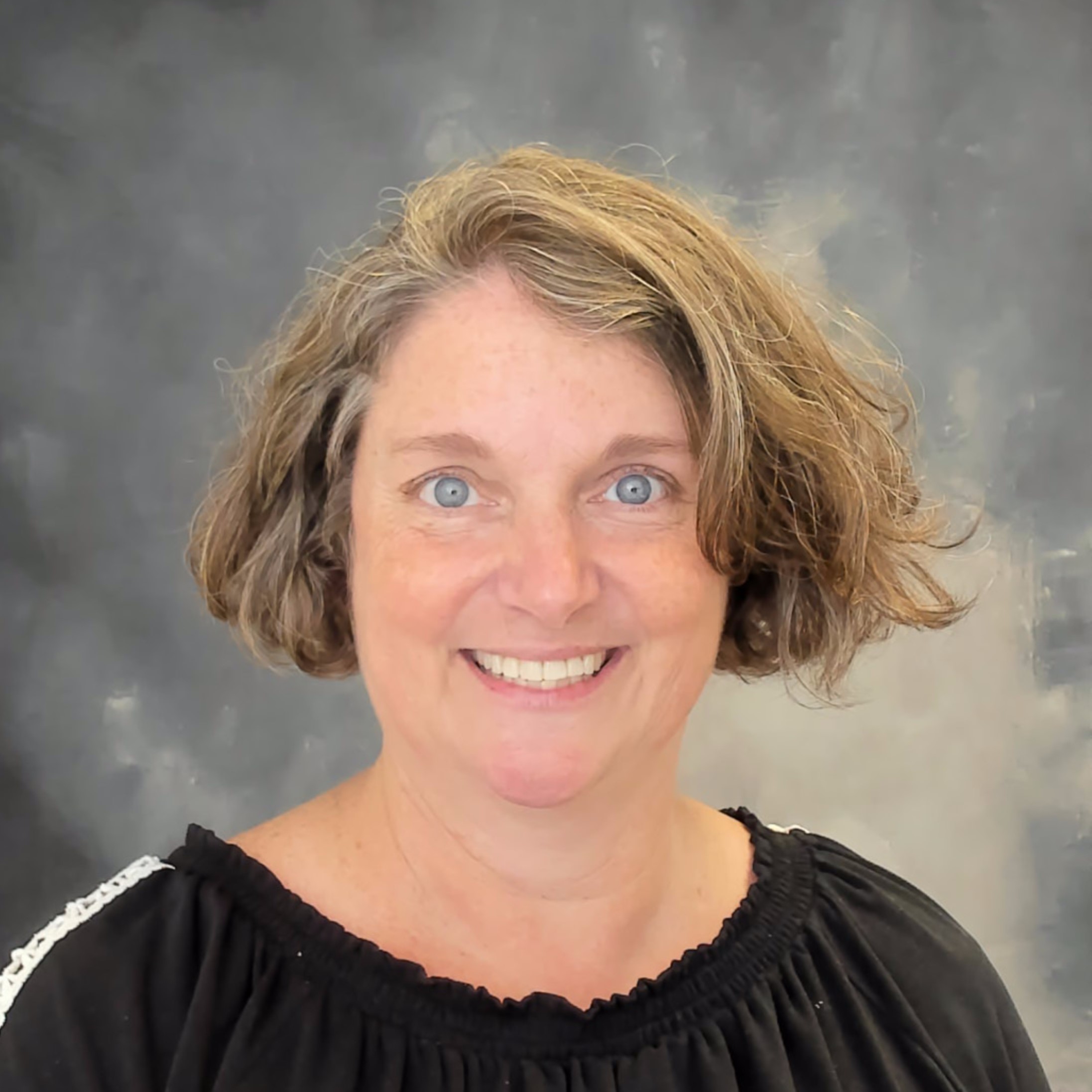 Christine Hausman is sitting in a portrait studio facing the camera. She is a middle aged white woman with greying short hair. Christine is wearing a black sweater.