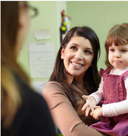 A woman with long brown hair holds her child while talking to a teacher