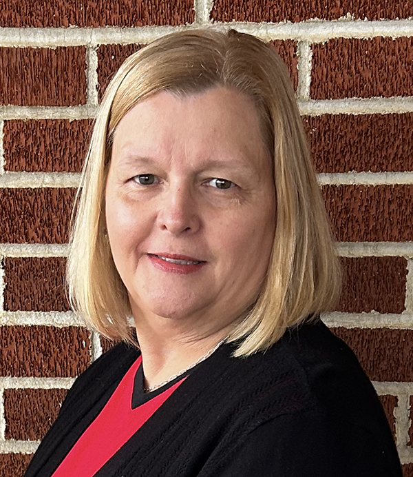 Headshot of a woman with shoulder length blonde hair wearing a red and black shot standing in front of a red brick wall.